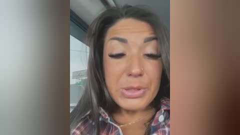 Media: A video of a smiling, light-skinned woman with long dark hair, wearing a plaid shirt, captured from the waist up inside a car.