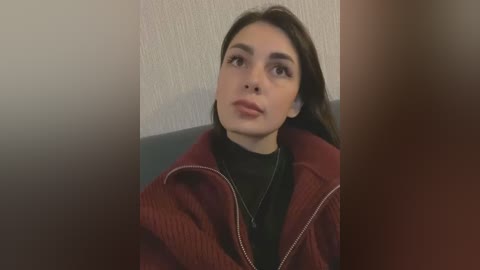 Media: Video of a young woman with olive skin, long dark hair, and a neutral expression, wearing a black turtleneck under a red quilted jacket. She sits against a textured beige wall.