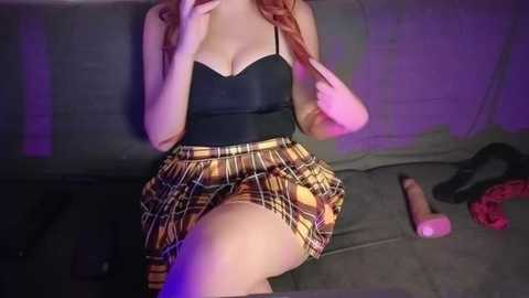 Media: Video of a fair-skinned woman with red hair, wearing a black bra and a plaid skirt, sitting on a grey couch, surrounded by sex toys and a red dress.