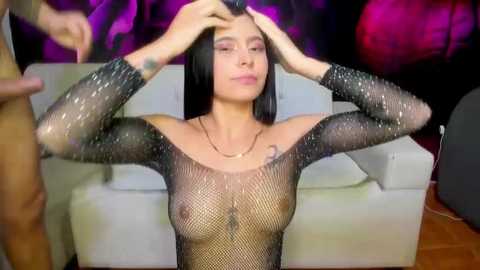 Media: Video of a slender woman with long black hair, wearing a sheer black mesh top, seated on a white couch, with purple and pink background lights.