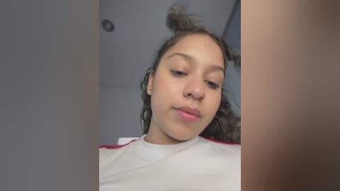 Media: Video of a young woman with light brown skin, curly dark hair, and a white shirt. She has a serene expression, looking down. The background is a simple, gray room with a ceiling light.