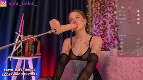 Media: A video of a light-skinned, red-haired woman with tattoos, wearing black lingerie and a large, realistic dildo, performing on a stage with flowers and a pink backdrop.