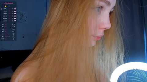 Media: Video of a young Caucasian woman with long, straight, blonde hair, seen in profile. She has pale skin and is topless. Background features a dimly lit room with a TV displaying a schedule, and a bright, round light in the foreground.