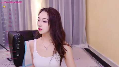 Media: Video of a young Asian woman with long dark hair, wearing a white tank top, sitting on a black gaming chair in a minimalist room with beige walls and gray curtains.