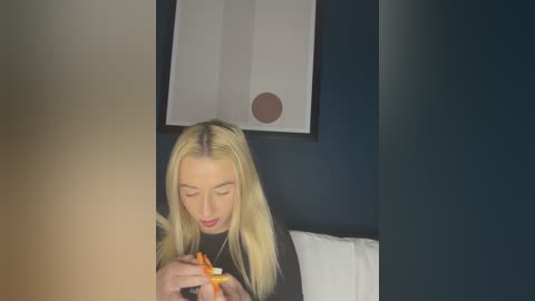 Media: A video of a blonde woman with fair skin, wearing a black shirt, focused on a cigarette, sitting on a bed against a dark wall with a modern abstract painting.