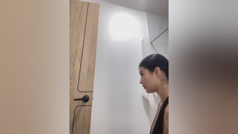 Media: Video of a young woman with medium brown skin and dark hair, wearing a black top, standing in a minimalist, white-tiled bathroom with a wooden door and a towel hanging on the wall.