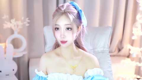 Media: Video of a young Asian woman with fair skin, light pink hair, and blue eyes, wearing a blue off-shoulder dress, sitting in a softly lit room with white curtains and a fluffy white pillow.