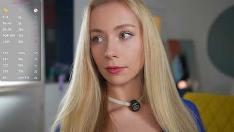 Media: Video of a young, light-skinned, blonde woman with long hair, wearing a blue top and a white choker, looking contemplative in a modern, softly lit room.