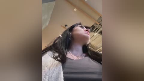 Media: A video of a young woman with long, dark hair, wearing a beige cardigan over a gray shirt, looking upwards in a dimly lit room with beige walls and a wooden ceiling.