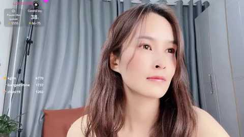 Media: Video of a young Asian woman with long, straight brown hair, fair skin, and light makeup, wearing a sleeveless top, standing in a modern room with gray curtains and a brown pillow.