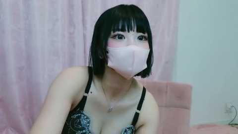 Media: Video of an Asian woman with a pale complexion, wearing a black lace bra and pink surgical mask, seated in front of a pink curtain background.