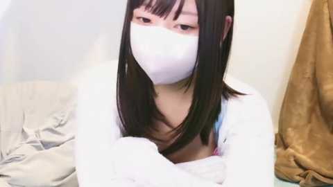 Media: A video of an Asian woman with long black hair, wearing a white face mask, white long-sleeve top, and blue bra, sitting on a bed with messy sheets, in a plain room.