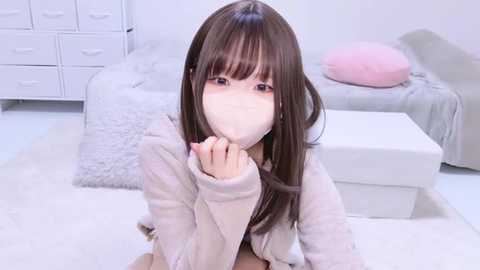 Media: Video of an Asian woman with straight, shoulder-length brown hair, wearing a white mask, a light pink jacket, and a white shirt, kneeling on a fluffy white carpet in a minimalist, white room with a bed and drawers.