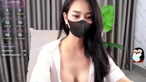 Media: Video of an Asian woman with long black hair, wearing a white blouse, black mask, and a penguin sticker in the corner. She sits in front of grey curtains and a green leafy plant.