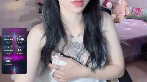 Media: Video of a young woman with long black hair, wearing a white lace bra, sitting in a pink bedroom with plush toys, displaying a smartphone game screen.