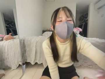 Media: Video of an Asian woman with long brown hair, wearing a yellow long-sleeve shirt, black shorts, and a light blue mask, sitting on the floor in a cluttered, dimly lit bedroom with an unmade bed and clothes scattered around.