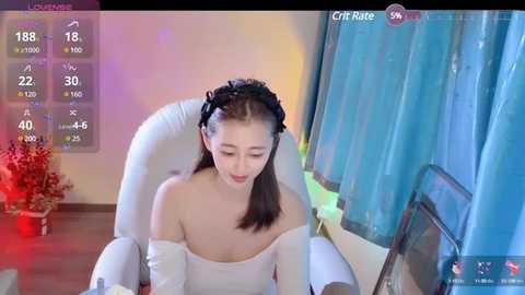 Media: A video of a fair-skinned East Asian woman with straight brown hair, wearing a white off-shoulder dress, seated in front of a blue curtain, with a computer screen showing a video chat interface.