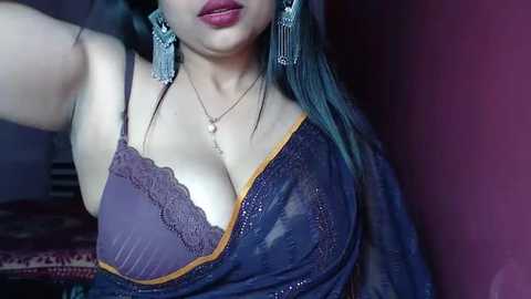 Media: Video of a woman with medium skin tone, wearing a purple lace bra and a sheer purple blouse, with long dark hair, large silver earrings, and a necklace, against a maroon background.