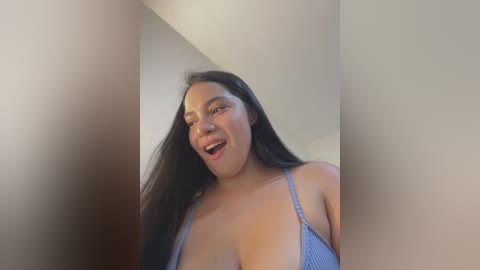 Media: Video of a smiling Latina woman with long black hair, wearing a blue ribbed tank top that reveals cleavage, standing in a dimly lit, beige-walled room.