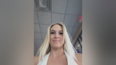 Media: Video of a smiling blonde woman with fair skin, wearing a white halter top, taken from a low angle. The background shows a gray ceiling with fluorescent lights and blurred objects, likely in an office or indoor setting.