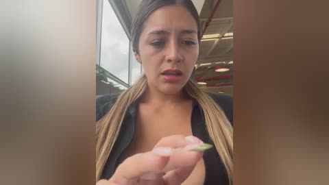 Media: Video of a young woman with long blonde hair, wearing a black top, holding a small green object, indoors near a glass window.