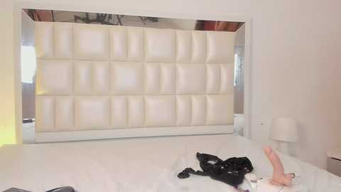 Media: Video of a modern bedroom with a large, white, tufted headboard against a wall. The bed features a white comforter, with black clothes scattered on it. Minimalist decor includes a white lamp on the right.