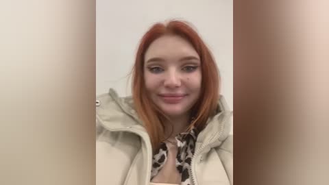 Media: Video of a fair-skinned young woman with long, straight, red hair, wearing a beige puffy jacket over a leopard print blouse. She smiles softly, with the background blurred.