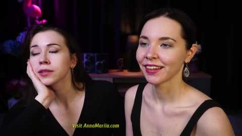 Media: Video of two women with fair skin and dark hair, smiling. One has a slender frame, the other has a fuller figure. They wear black dresses, and the background is dimly lit with purple and pink hues.