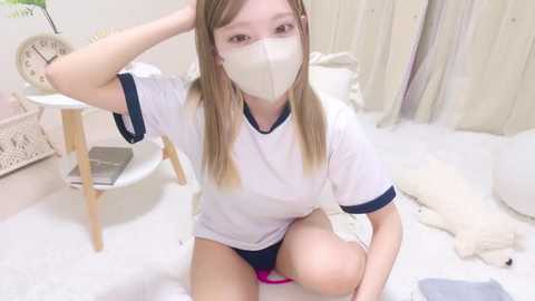 Media: Video of a young East Asian woman with light skin and long, straight, light brown hair. She's wearing a white T-shirt, blue panties, and a face mask. The room is minimalist, with white decor and a plush toy.