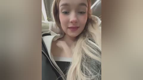 Media: Video of a young woman with light skin and long blonde hair, wearing a black jacket with a white collar, sitting in a car with beige seats.