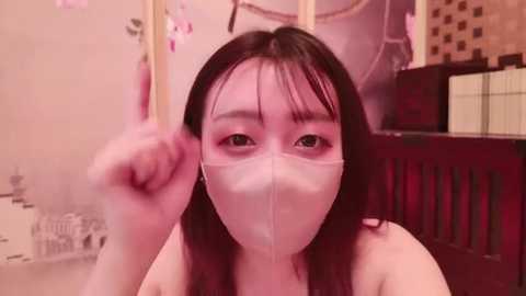 Media: A video of an East Asian woman with long black hair and fair skin, wearing a pink surgical mask, making a fist in a room with cherry blossom decorations and a wooden chair in the background.