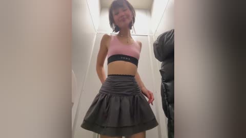 Media: Video of a slender Asian woman with shoulder-length dark hair, wearing a pink sports bra and a gray pleated skirt, standing in a narrow hallway with white walls.