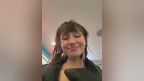 Media: Video of a smiling young woman with straight, shoulder-length brown hair, wearing a black garment, standing in a brightly lit room with modern decor.