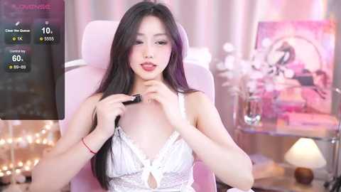 Media: Video of a young Asian woman with long black hair, wearing a white lace bra, applying makeup in a pink chair; background features soft lighting, flowers, and a framed photo.