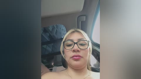 Media: Video of a young, plus-sized Latina woman with shoulder-length blonde hair, wearing black-rimmed glasses, pink lipstick, and a white top, sitting in a car, with a blue patterned seat behind her.