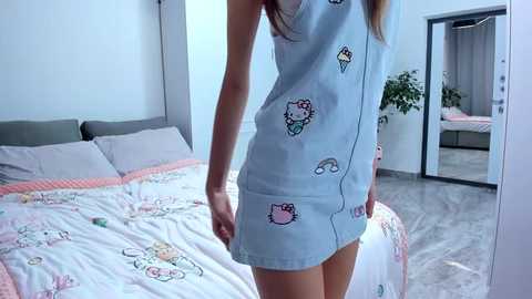 Media: Video of a slender woman in a light blue, sleeveless Hello Kitty nightgown with cartoon characters, standing in a modern bedroom with a white bed, pink sheets, and a potted plant.