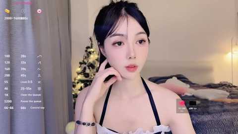 Media: Video of a young Asian woman with fair skin and long black hair, wearing a black and white halter top, sitting in a cozy bedroom with a lit Christmas tree.