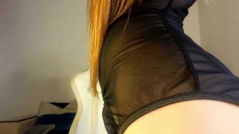 Media: Video of a close-up of a woman's upper thigh and hip in a sheer black bodysuit, revealing smooth, light skin. Her long, straight, reddish-brown hair cascades down her back. Background shows a white chair and a dark cushion.