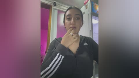 Media: Video of a young woman with medium skin tone, dark hair, and a neutral expression, wearing a black Adidas hoodie, standing in a brightly lit room with a pink door and a refrigerator in the background.