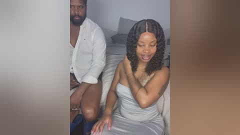 Media: Video of a Black couple in a dimly lit bedroom: man in white shirt, shorts, sitting; woman in strapless grey dress, sitting, hands touching her neck, looking down.