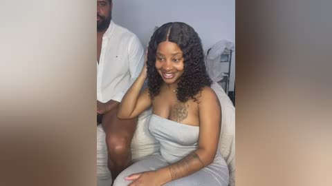 Media: Video of a Black woman with curly hair in a strapless white dress, sitting on a couch with a man behind her in a white shirt, smiling in a simple room with minimal decor.