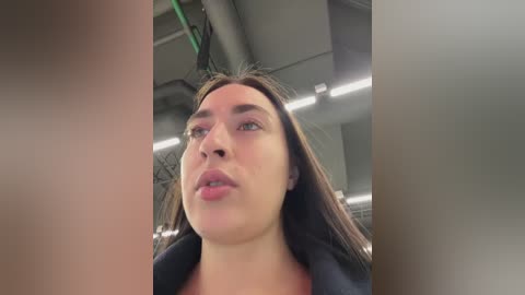 Media: Video of a light-skinned woman with long brown hair, wearing a black jacket, looking introspective in a modern, industrial setting with metallic ceiling pipes and green cables.