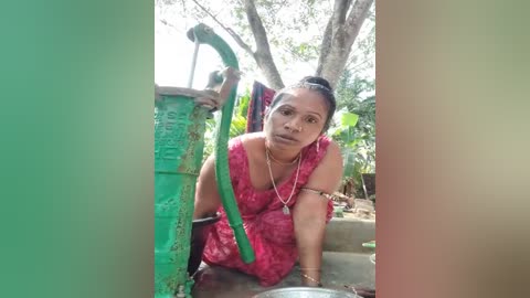 Media: Video of a South Asian woman in a pink floral dress, leaning on a green metal pipe with a water pump in a rural setting.