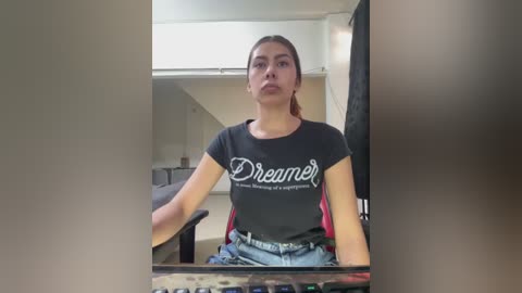 Media: Video of a young woman with medium skin tone, long brown hair, wearing a black \"Dream\" T-shirt and blue jeans, sitting in an office chair, with a blurred background of office furniture.