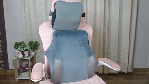 Media: Video of a pink gaming chair with a blue cushion on its backrest, positioned in a room with beige curtains and wooden floor, featuring a small plant and shelves with decorative items in the background.