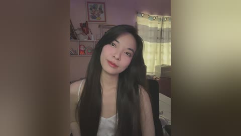 Media: Video of a young Asian woman with long black hair, light skin, wearing a white tank top, smiling in a softly lit bedroom with wall-mounted shelves and a window with white curtains.