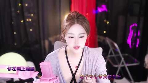 Media: Video of a young Asian woman with fair skin and brown hair in a bun, wearing a black bra and a light grey cardigan, sitting at a table with pink cake, balloons, and a green candle.