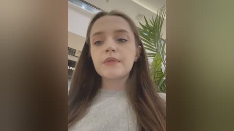 Media: Video of a young Caucasian woman with long brown hair, wearing a light-colored sweater, standing indoors with green plants in the background.