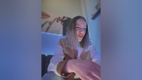 Media: Video of a smiling, light-skinned woman with shoulder-length, straight blonde hair, wearing a beige and white sweater, sitting on a dark sofa. Background features abstract art with red lips.