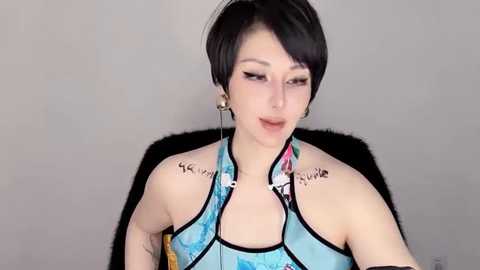 Media: Video of a pale-skinned, androgynous person with short black hair and large hoop earrings, wearing a blue halter top, seated indoors against a plain gray background.